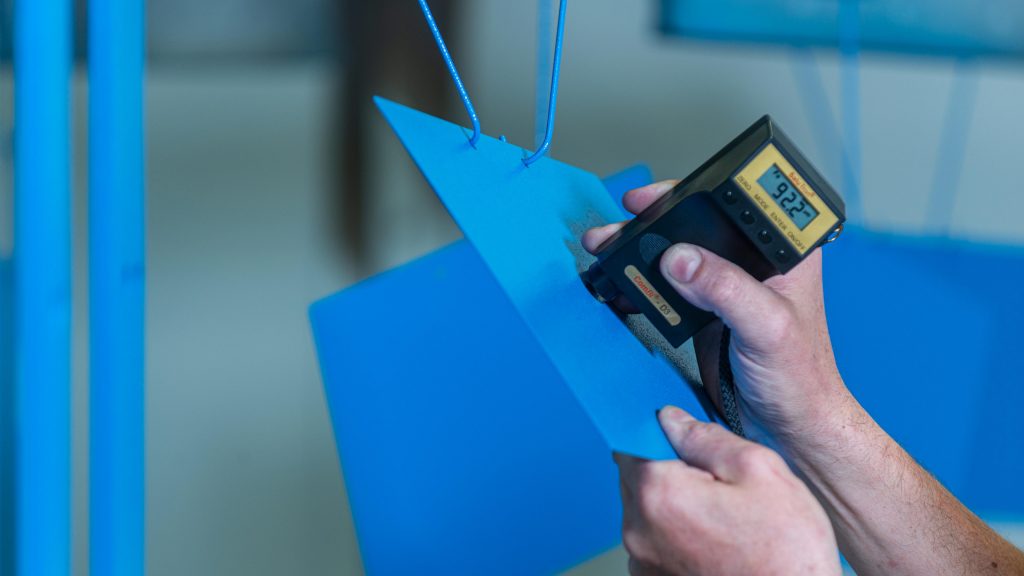 Farbschichtprüfung bei JORDAN: Hände setzen ein Messgerät auf ein mit blauer Farbe beschichtetes Blech